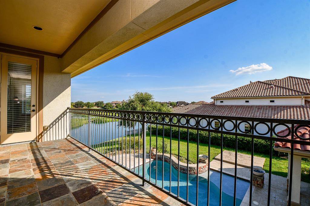 This is a spacious, covered balcony with a picturesque view overlooking a serene backyard pond and a neighboring house with a tiled roof. The balcony features attractive stone flooring and a decorative wrought-iron railing, perfect for outdoor relaxation and entertainment.