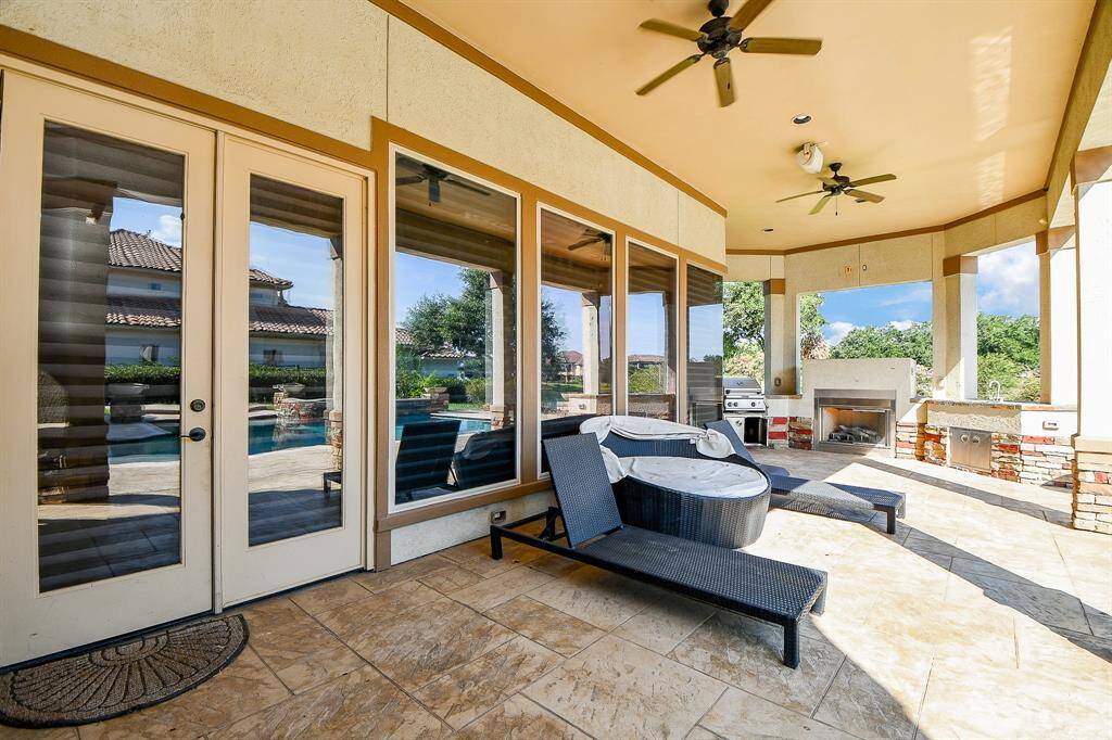 This is a spacious covered patio with tile flooring, featuring an outdoor kitchen with grill and fireplace, ceiling fans for comfort, and sliding glass doors leading into the home. It overlooks a pool area, suggesting a luxurious outdoor living space.