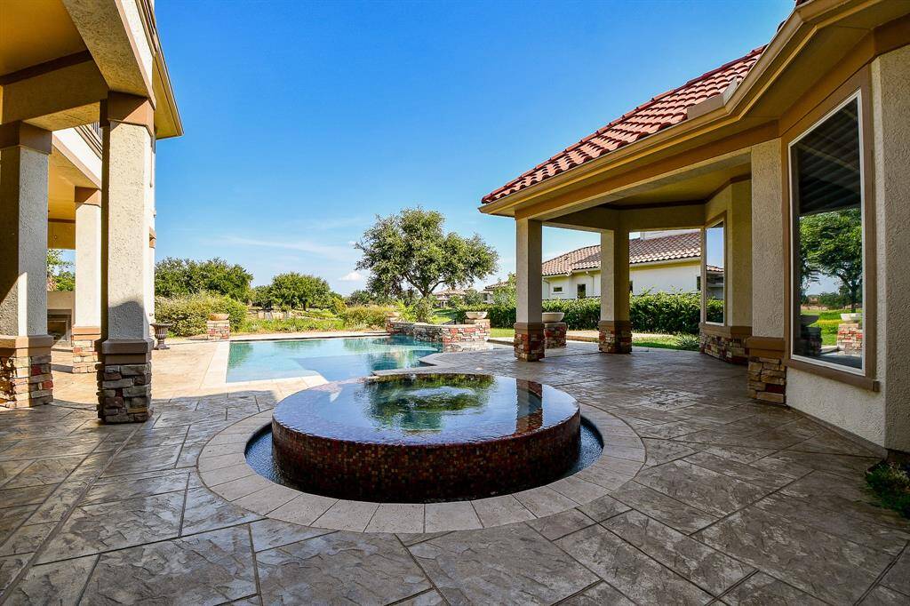 This image showcases a spacious outdoor patio with a luxurious pool and an integrated circular spa, all set within a private, landscaped backyard. The covered patio area features classic columns and elegant tile flooring, perfect for outdoor entertaining.