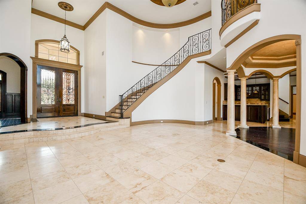 This is a spacious entryway with high ceilings, an elegant staircase, and travertine flooring. Classic architectural details include columns and archways, leading to an inviting living space beyond.