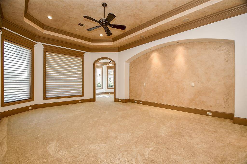 This is a spacious, primary bedroom suite with carpeted flooring, large windows with blinds, tray ceilings with recessed lighting, and a ceiling fan. It has a neutral color scheme and an arched doorway leading to another room.