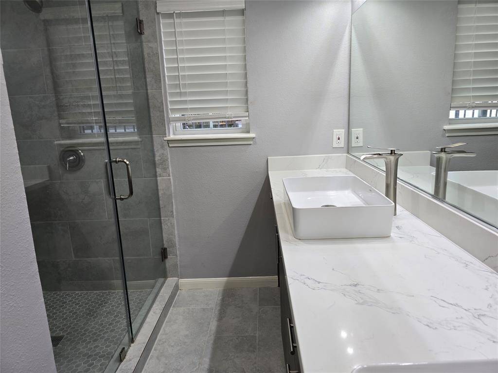 This modern bathroom features a glass-enclosed shower with gray tile, a stylish white vessel sink on a new countertop, and ample natural light from a new window with blinds.