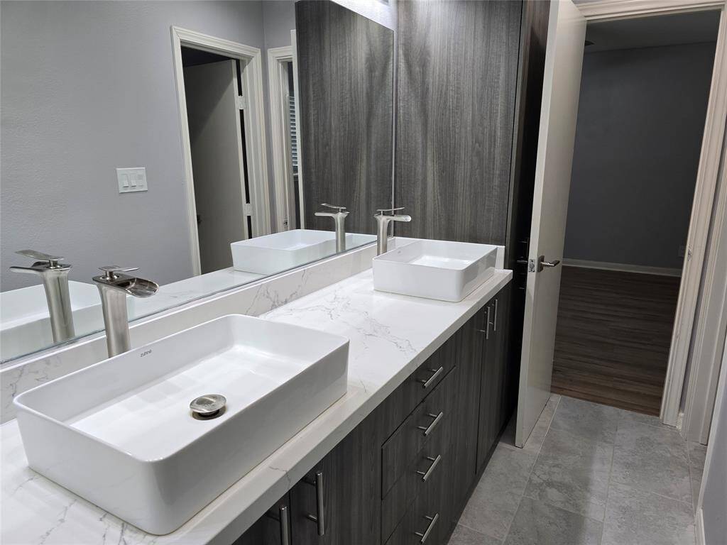 This bathroom features a modern double vessel sinks, new faucets, and large mirrors. The dark cabinetry complements the light tile flooring, creating a clean and contemporary look.