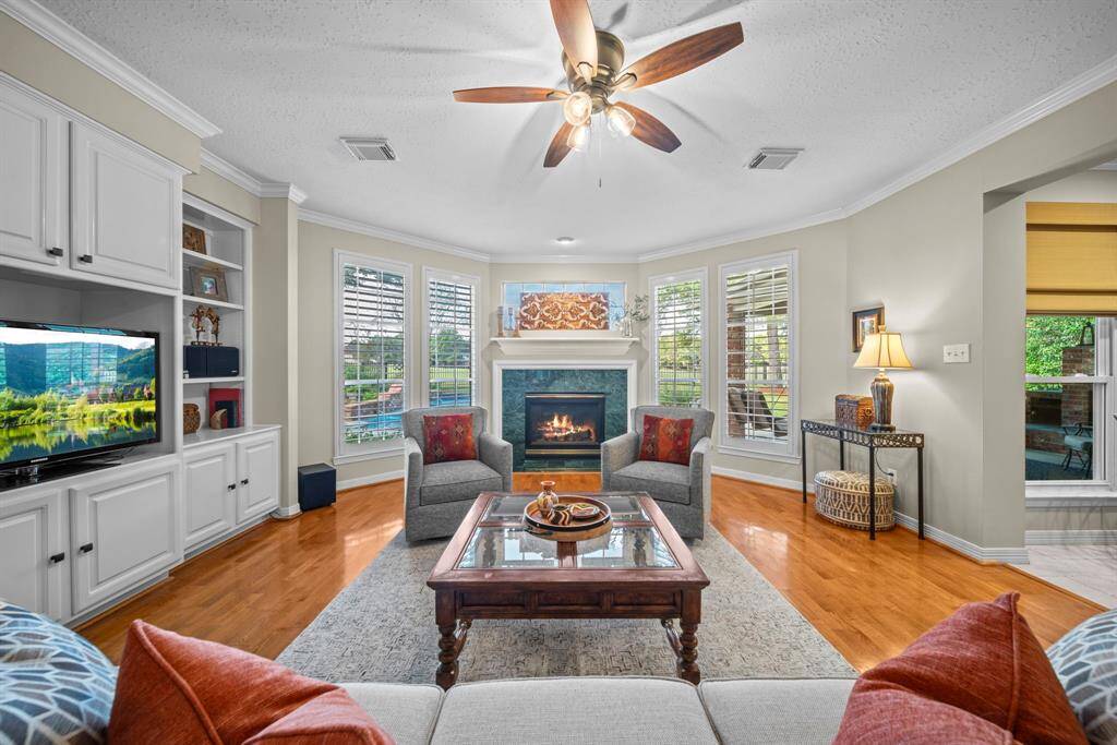 The spacious living area boasts classic wood flooring, crown molding, a ceiling fan, built-ins, a cozy fireplace, and recessed lighting perfect for showcasing your favorite artwork