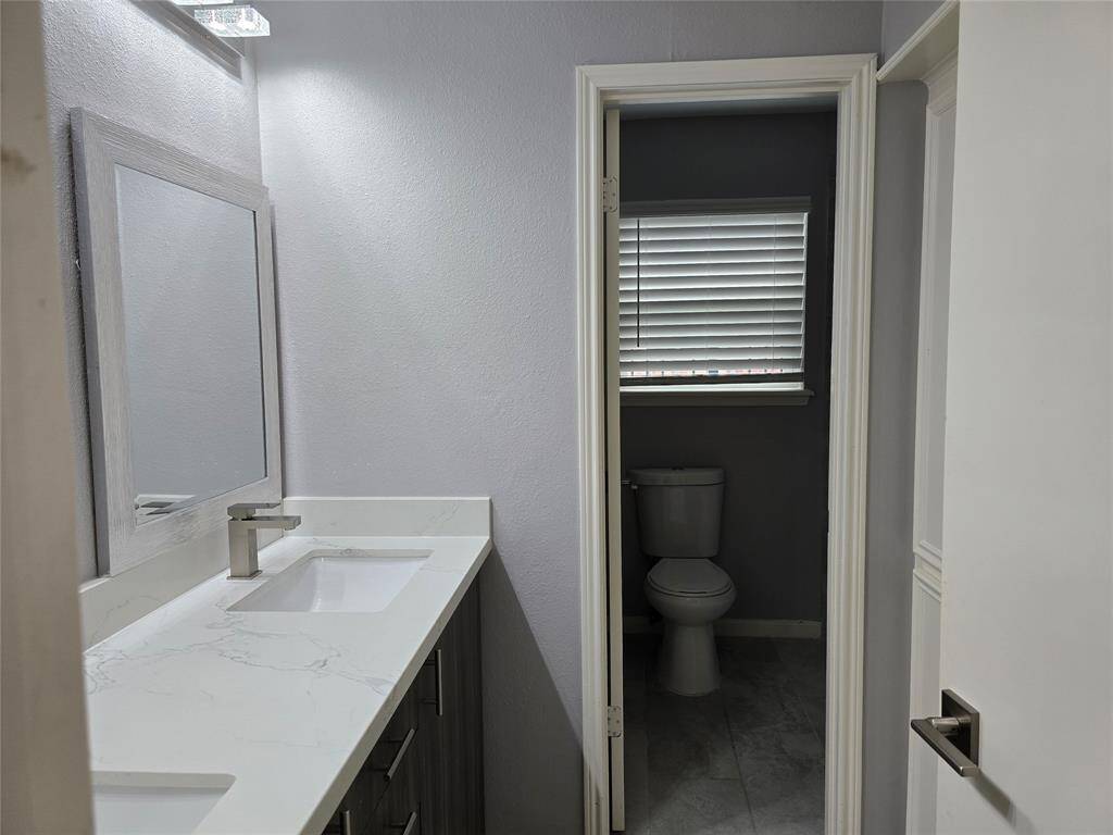 This modern second full bathroom features a new countertop with double under-mounted sinks and contemporary faucets. A separate toilet area features a window with blinds, enhancing natural light.