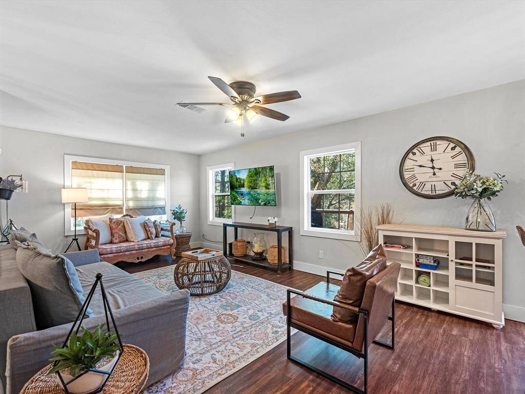 This inviting living room features hardwood floors, a ceiling fan, and abundant natural light from large windows. It includes comfortable seating with a mix of modern and traditional styles, a wall-mounted TV, and a decorative wall clock. The space is accented with a neutral color palette and tasteful decor.