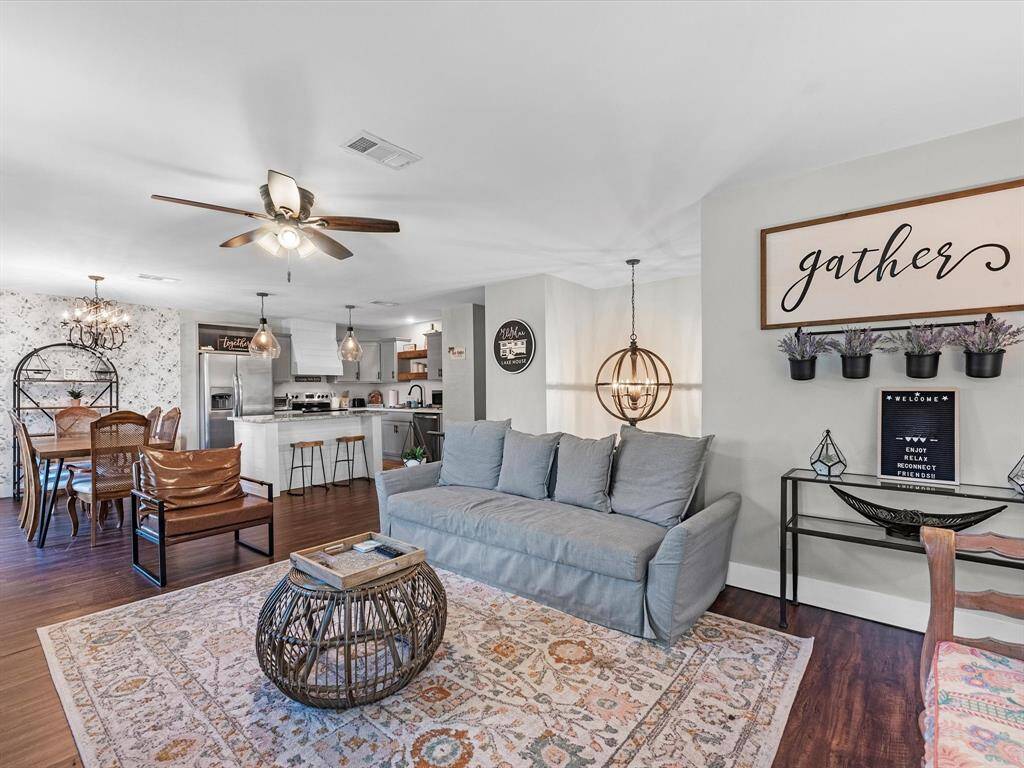 This inviting open-concept living space features a cozy seating area with a modern gray sofa and stylish lighting. Adjacent is a dining area with a wooden table and chairs, accented by a chic chandelier. The kitchen boasts stainless steel appliances, a breakfast bar, and pendant lights. The room is decorated in warm tones with charming wall art, creating a welcoming atmosphere.