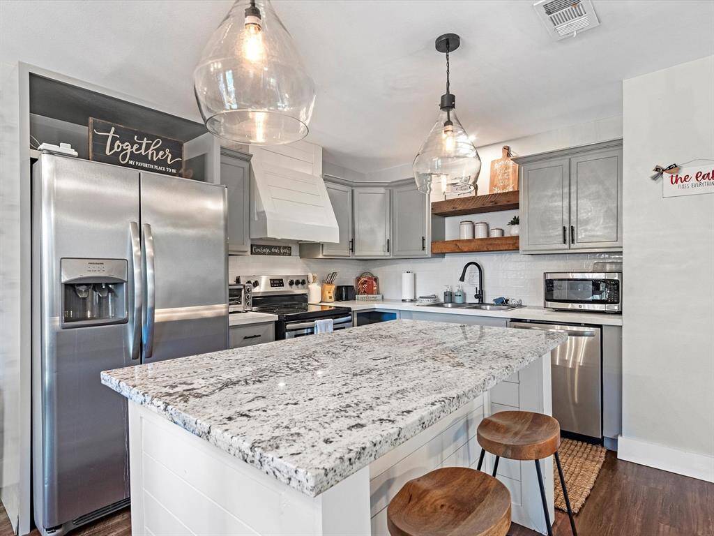 This modern kitchen features a spacious granite island with seating, stainless steel appliances, and stylish pendant lighting. It includes ample cabinetry for storage and open wooden shelves for added charm. The layout is both functional and inviting, perfect for cooking and entertaining.