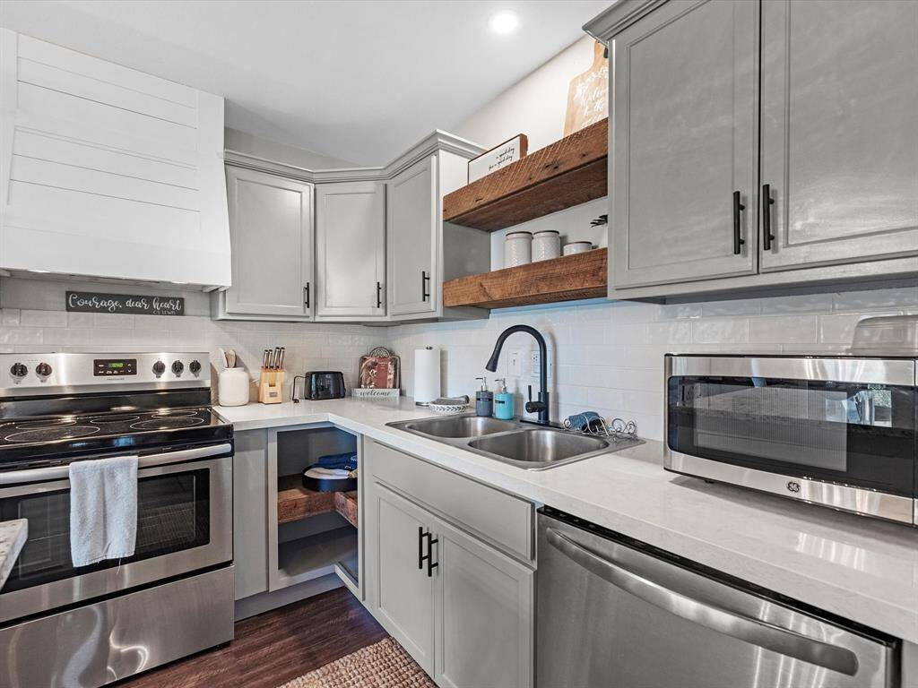 This kitchen features modern stainless steel appliances, light gray cabinetry, and open wooden shelves. It has a sleek, clean design with a subway tile backsplash and a functional layout, perfect for home cooking.