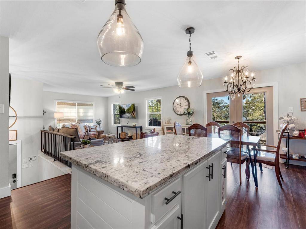 Bright, open-concept living space featuring a modern kitchen with a large island, granite countertops, and stylish pendant lighting. The adjacent dining area has a chandelier and large windows, offering a view of the greenery outside. The living area is cozy with ample seating and natural light.