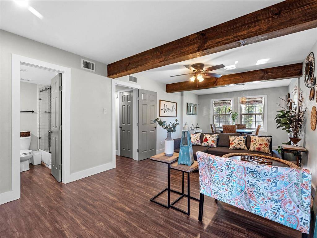 This inviting living space features exposed wooden beams and a ceiling fan, offering a cozy atmosphere. The room is bright with natural light from large windows, and it connects to a dining area. The open layout includes stylish decor and access to a modern bathroom.
