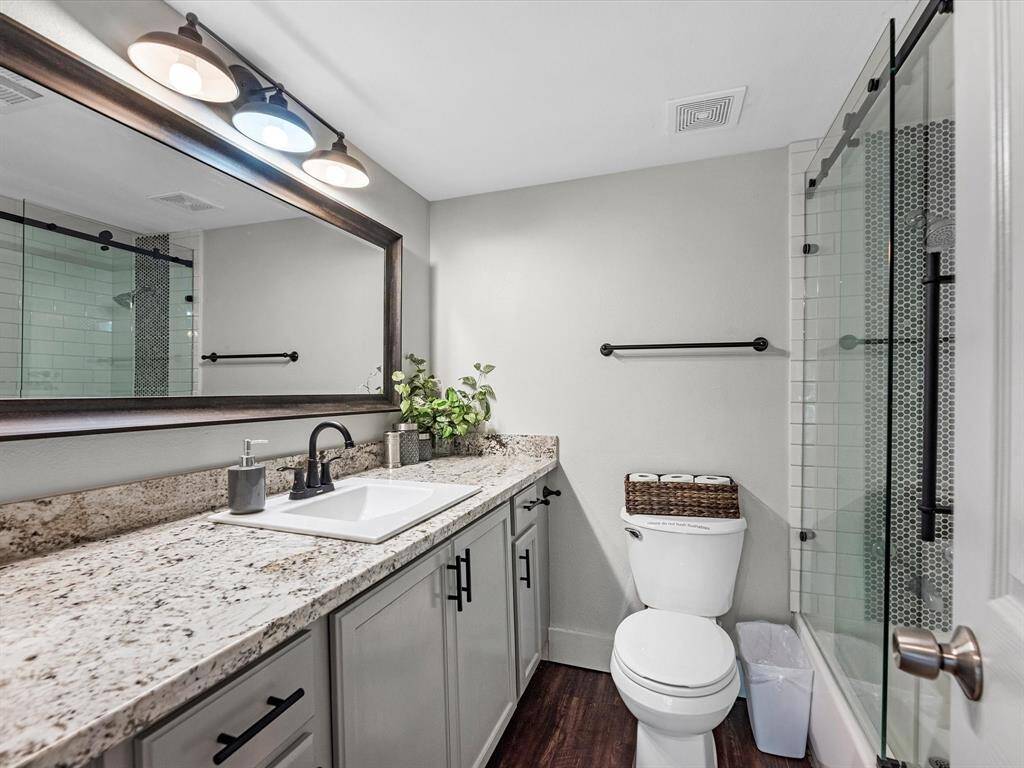 This bathroom features a modern design with a granite countertop, under-mount sink, and stylish fixtures. It includes a large mirror with overhead lighting, a glass-enclosed shower, and wood-look flooring for a warm touch.