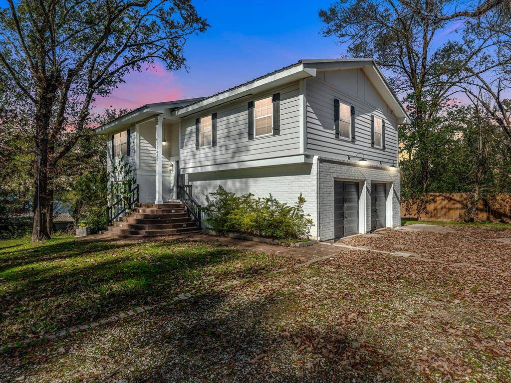 This charming two-story home features a white exterior with black shutters and a welcoming front porch with steps. It includes a two-car garage and is surrounded by mature trees, offering a serene and private setting. The driveway and yard are spacious, perfect for outdoor activities.