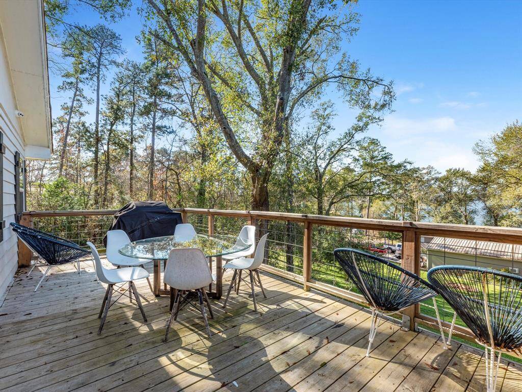 A spacious wooden deck with a scenic view of trees and water. It features modern outdoor furniture, including a glass-top table, chairs, and a grill, perfect for relaxing or entertaining.
