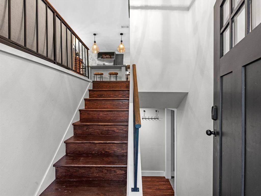This photo features a modern entryway with dark wood stairs leading to an upper level. The space is well-lit with stylish pendant lights, and there's a view of a cozy kitchen area with a breakfast bar and stools. The design is clean and contemporary, with a welcoming atmosphere.
