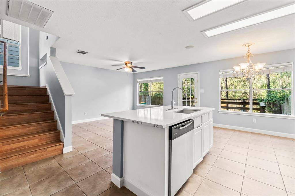 Bright and spacious kitchen and dining area with large windows, tile flooring, and a central island with a sink. Features a modern chandelier, ceiling fan, and adjacent to wooden staircase leading to the second floor.