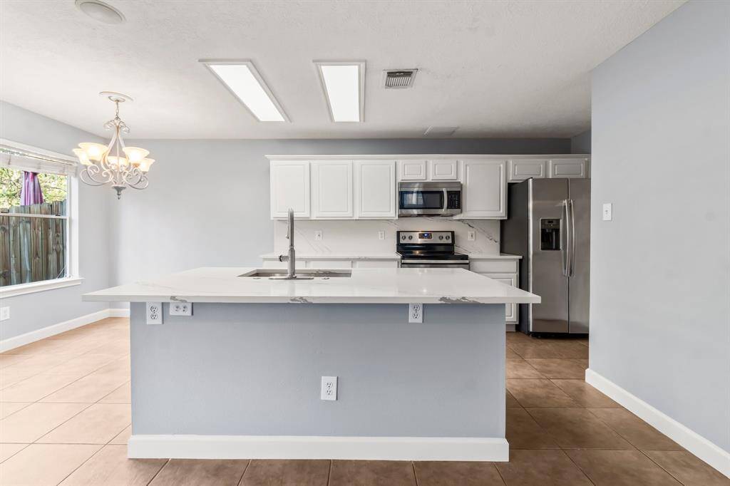 This kitchen features modern stainless steel appliances, white cabinetry, and marble-patterned countertops. It includes a bright dining area with large windows and tile flooring, enhanced by a stylish chandelier and ample natural light.