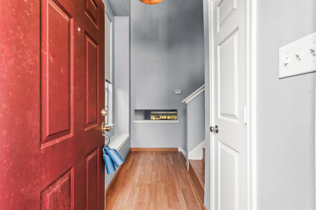 The photo shows a welcoming entryway with a red door leading to a hallway with light wood flooring. There's a small window ledge and a staircase to the right. The walls are painted a soft gray, adding a modern touch.
