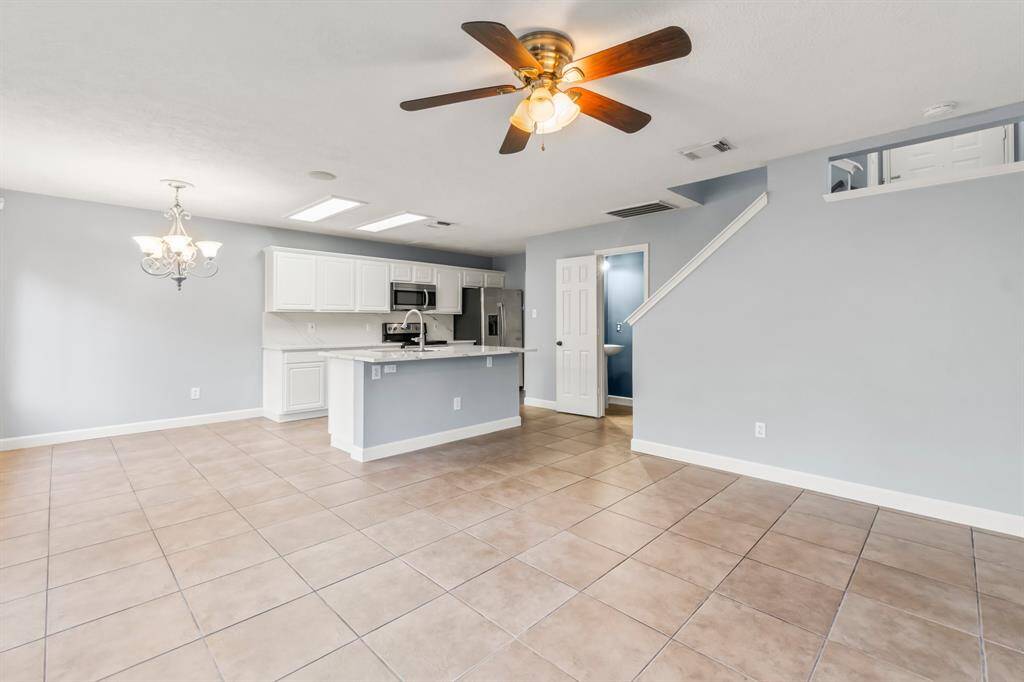 This photo showcases a spacious, open-concept kitchen and living area with tiled floors, light gray walls, and a ceiling fan. The kitchen features white cabinets, a modern stainless steel microwave, and a central island. A staircase and a light fixture add to the room's appeal.