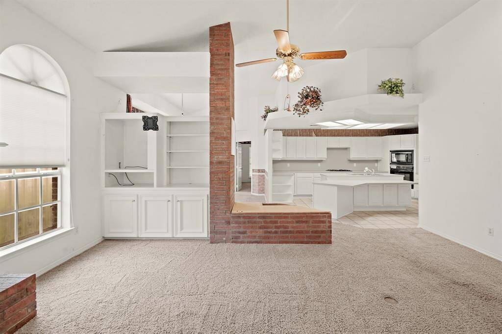 This photo showcases a spacious, open-concept living area with high ceilings. The room features a large window with natural light, a brick divider, built-in shelving, and a ceiling fan. The adjacent kitchen is equipped with white cabinetry, an island, and modern appliances, offering a bright and airy feel.