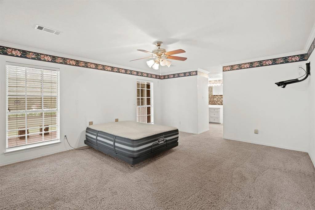 This spacious bedroom features cozy carpeting, a ceiling fan, and large windows for natural light. The room includes a floral border detail and an adjacent nook area leading to a bathroom. Ideal for creating a comfortable retreat.