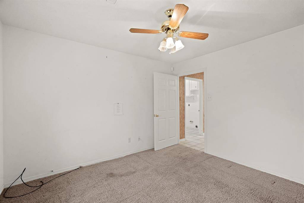 This is a clean, simple bedroom with neutral walls and carpet. It features a ceiling fan and a doorway leading to the laundry area. The space is well-lit and ready for personalization.