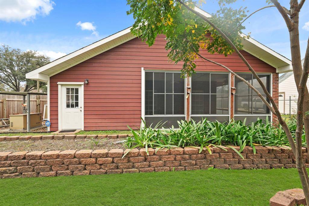 Charming backyard view of a red house with a screened porch, surrounded by lush greenery and a neat stone retaining wall. Perfect for enjoying outdoor relaxation.