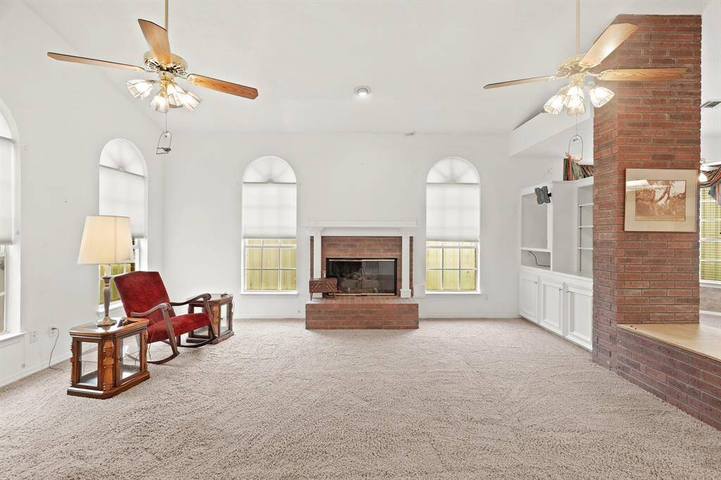 This spacious living room features a cozy brick fireplace, large windows for natural light, ceiling fans, and built-in shelving. The neutral carpet and walls create a warm and inviting atmosphere.