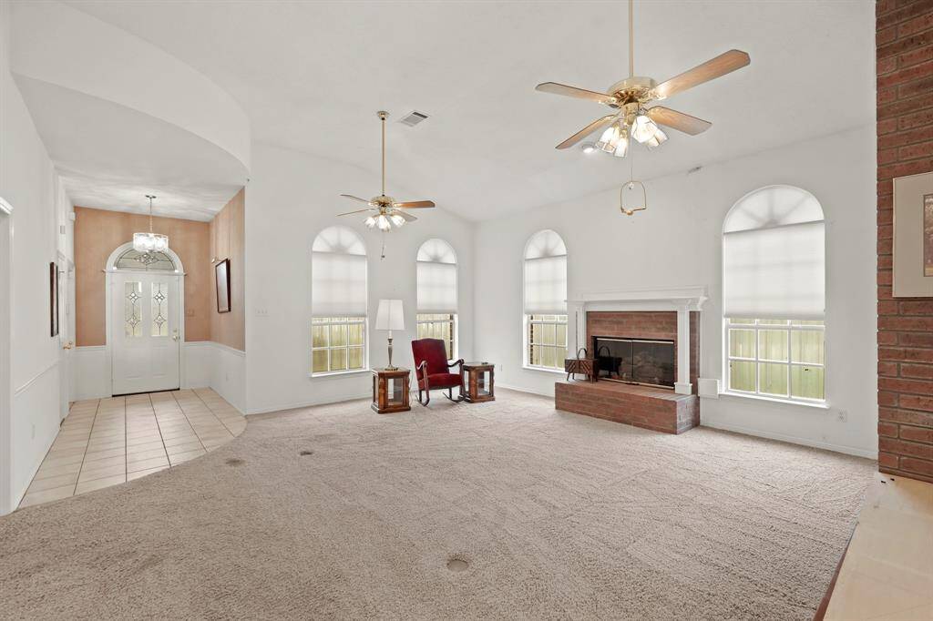 This spacious living room features high ceilings, large arched windows for natural light, and a cozy fireplace. The neutral carpeting and ceiling fans add comfort, while the entryway opens to a tiled foyer with a chandelier.