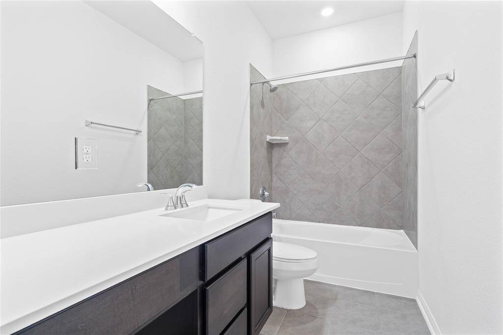 Secondary bath features tile flooring, bath/shower combo with tile surround, wood cabinets, beautiful light countertops, mirror, dark, sleek fixtures and modern finishes.
