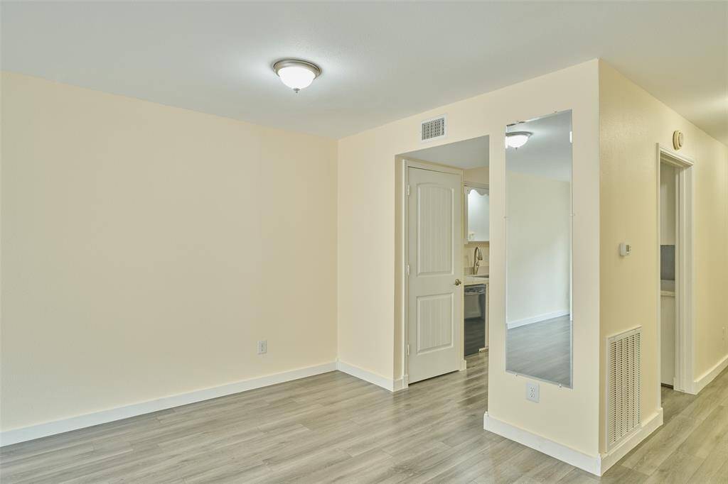 This photo shows a bright, open room with light-colored walls and sleek wood flooring. It features recessed lighting, a doorway leading to a kitchen with visible appliances, and a hallway, creating a welcoming atmosphere.