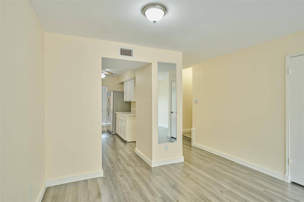 This photo showcases a bright, open space with light-colored walls and flooring, leading into a kitchen area. The kitchen features white cabinetry and stainless steel appliances, creating a modern and inviting atmosphere.