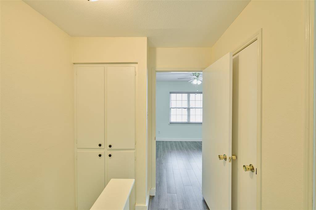 This photo shows a hallway with built-in storage cabinets and a doorway leading to a bright room with a large window and wood flooring. The space is well-lit and has a clean, neutral aesthetic.