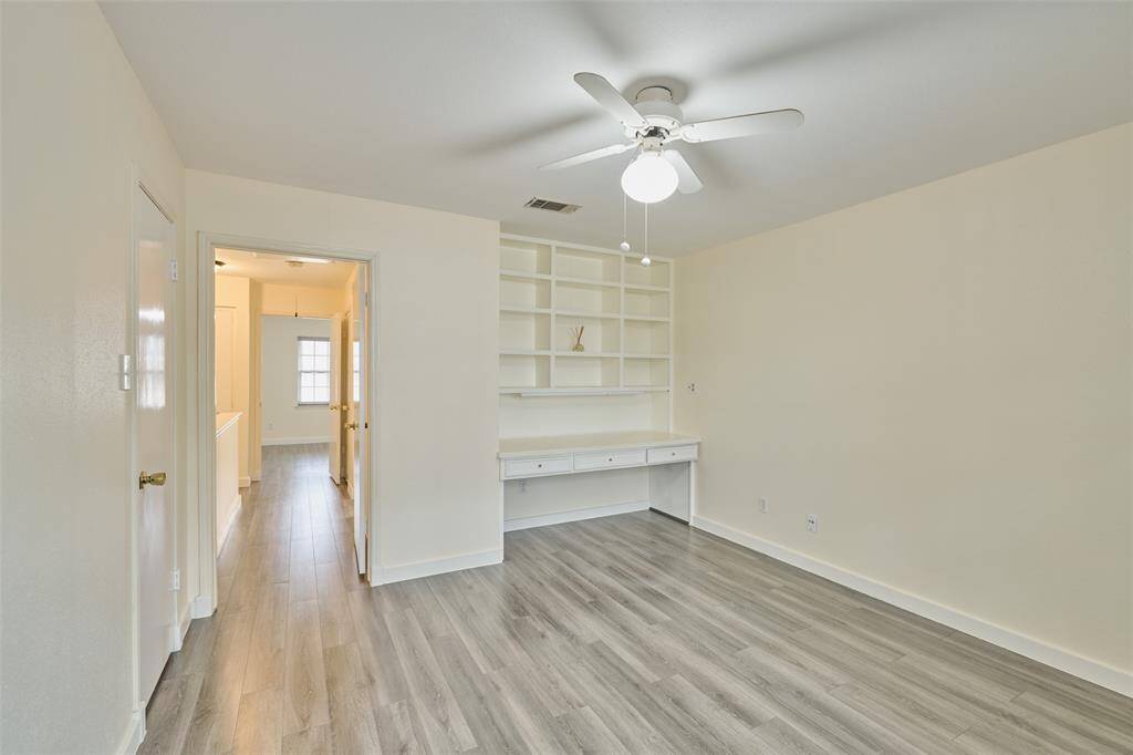 Bright and inviting room with modern gray laminate flooring and fresh neutral paint. Features include a ceiling fan, built-in shelving, and a desk area, offering functionality and style.