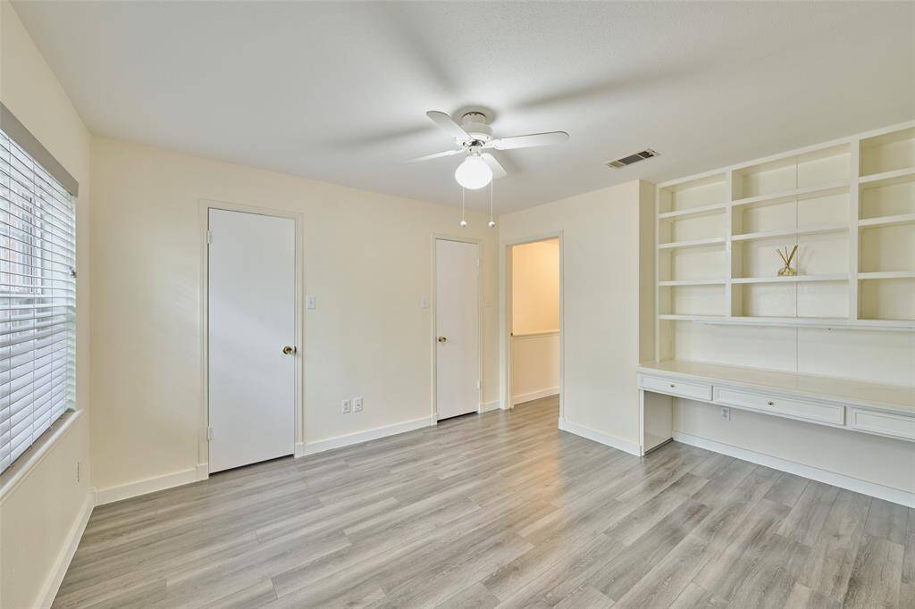 Bright room with light wood flooring and a ceiling fan. It features built-in shelves and a desk, ideal for an office or study space. Natural light streams through the window with blinds, enhancing the spacious feel.