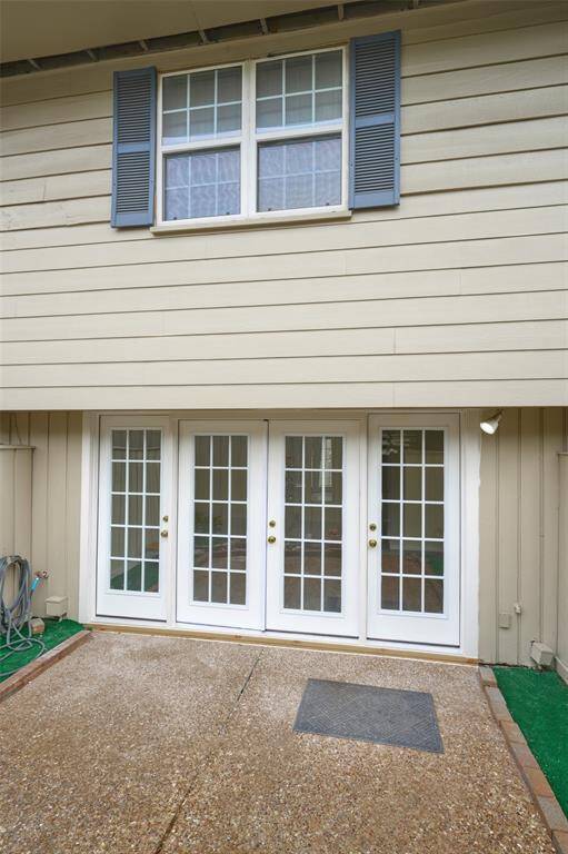This photo showcases a home's exterior with double French doors leading to a patio. The area features a textured concrete surface and is bordered by a small strip of artificial grass. Above, there are windows with blue shutters, adding a touch of charm.