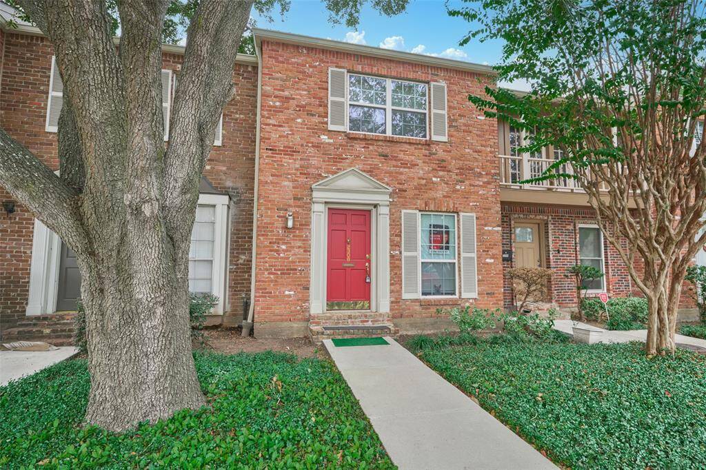Charming two-story brick townhouse with a striking red door, elegant white window frames, and lush landscaping featuring a large tree and vibrant ground cover.