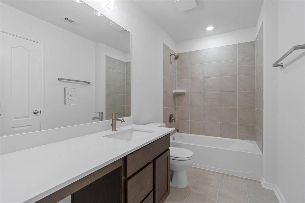 Secondary bath features tile flooring, bath/shower combo with tile surround, stained wood cabinets, beautiful light countertops, mirror, dark, sleek fixtures and modern finishes.