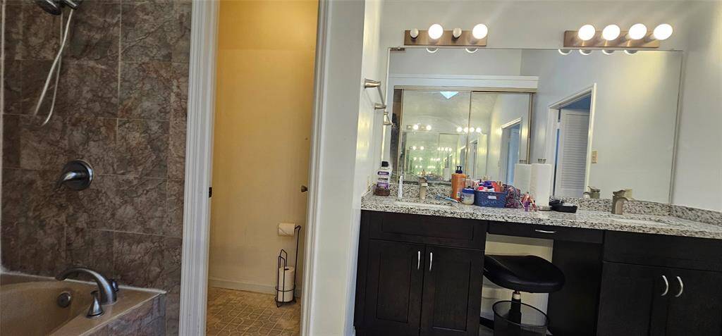 Primary bathroom featuring a granite countertop with dual sinks, a large mirror with ample lighting, and there's also a small seating area at the vanity.