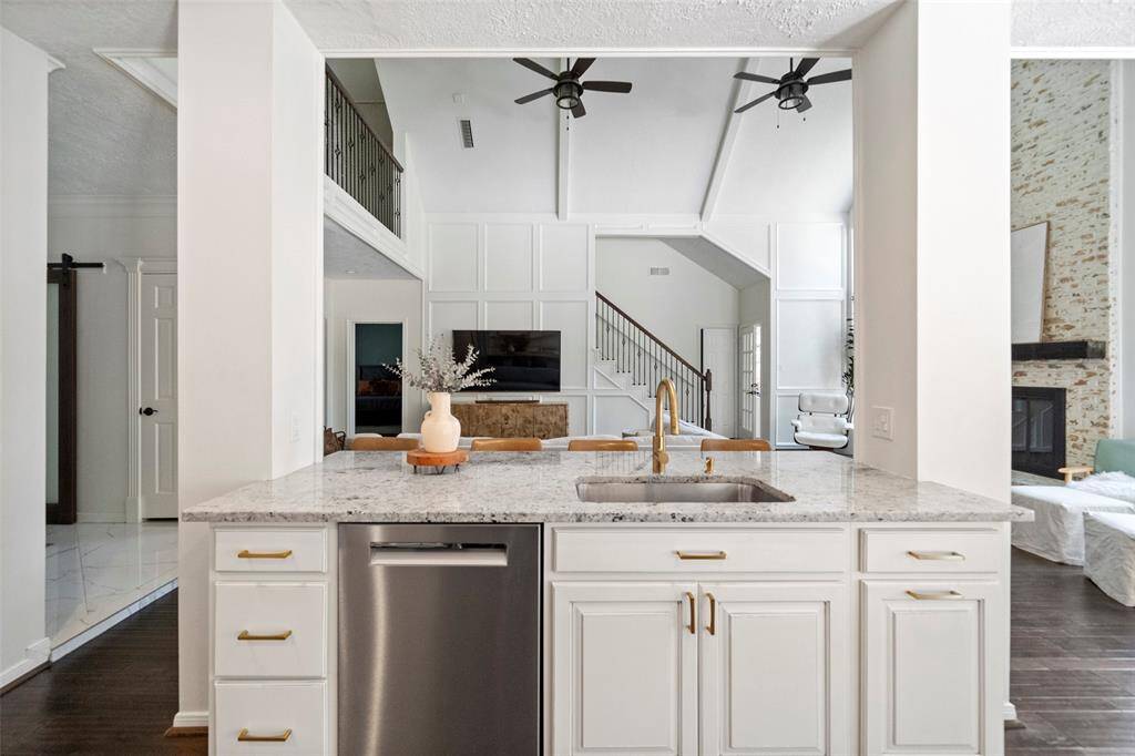 This photo showcases a modern, open-concept kitchen with a granite countertop, stainless steel dishwasher, and elegant fixtures. It opens into a bright living area with high ceilings, exposed beams, and a cozy fireplace, offering a spacious and inviting atmosphere.