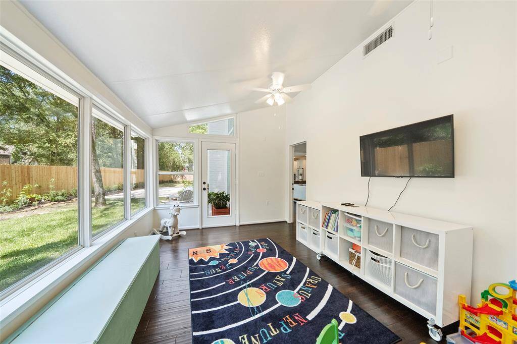 This bright sunroom features large windows overlooking a lush backyard, dark wood flooring, a ceiling fan, and ample storage with a TV mounted on the wall. It's a perfect space for relaxation or a play area.