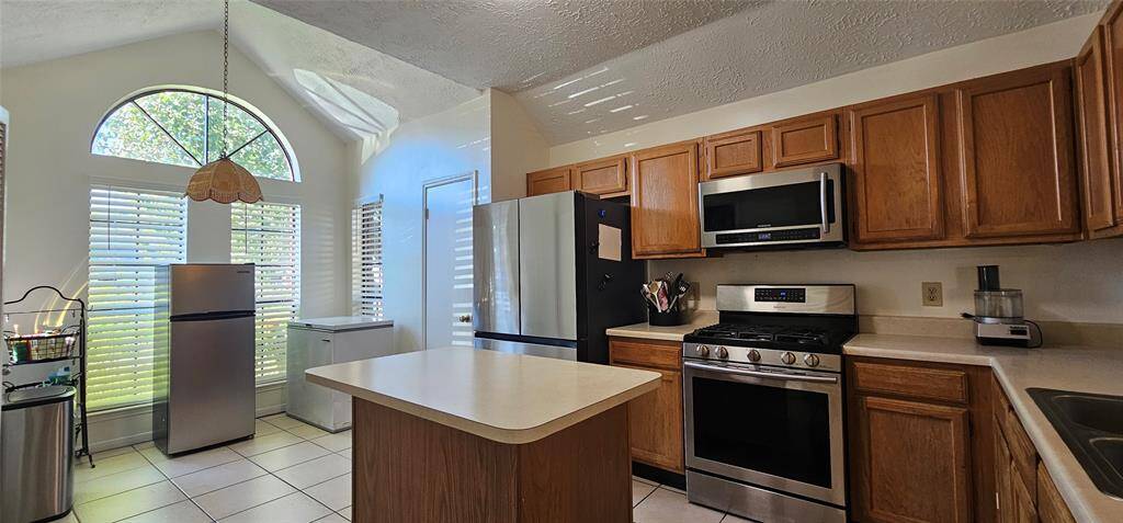 This kitchen features ample natural light with large windows, a central island, stainless steel appliances, and warm wood cabinetry. The space is open and inviting, ideal for cooking and entertaining.