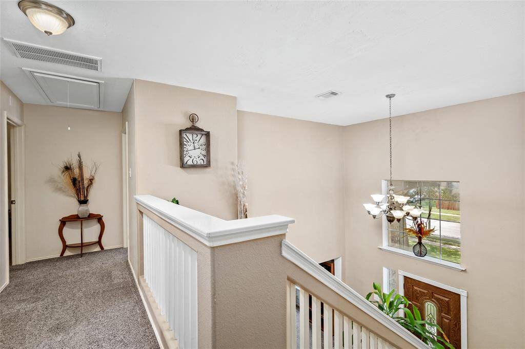 The second floor hallway has neutral wall colors, carpeted flooring, and a classic chandelier. The space includes a wooden railing and a window offering natural light and a view of the outside.