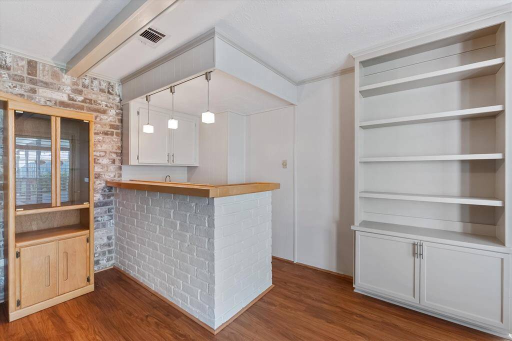 This home features a cozy nook with a small wet bar area, highlighted by pendant lighting and a white brick accent. Adjacent are built-in shelves and a cabinet, offering ample storage and display options. The woodwork on the bar was hand done by the owner. The space has a warm ambiance with a mix of painted brick and wood flooring.