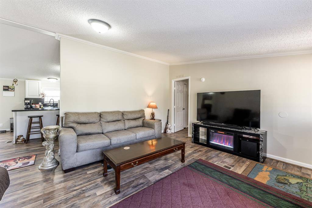 Cozy living room with wood flooring, featuring a large TV on a stand with an electric fireplace. There's a comfortable gray sofa, a wooden coffee table, and a small table lamp. The space is open to the kitchen, which includes a breakfast bar with stools.
