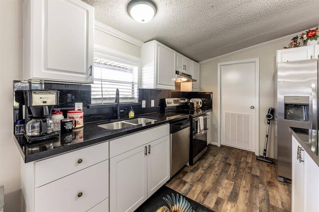 This kitchen features modern stainless steel appliances, sleek black countertops, and white cabinetry. It has a cozy layout with a window above the sink providing natural light, and stylish wood-look flooring.