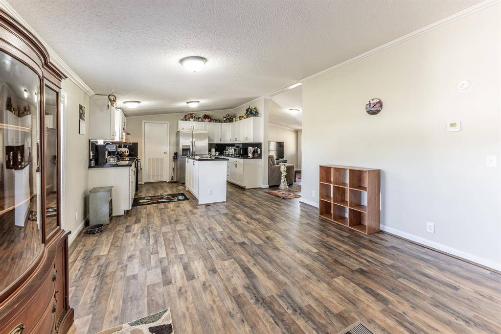 This photo showcases a spacious open-plan living area with a modern kitchen. The room features wood-style flooring, ample lighting, and neutral walls. The kitchen includes white cabinetry, a central island, and stainless steel appliances, creating a welcoming and functional space.