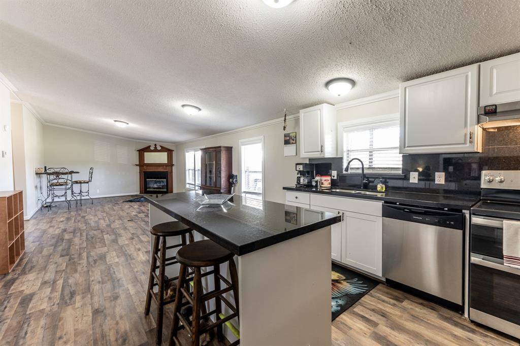 This open-concept kitchen and living area features sleek stainless steel appliances, white cabinetry, and a modern island with seating. The cozy living space includes a fireplace, and large windows provide ample natural light. The flooring is a warm wood tone, enhancing the inviting atmosphere.
