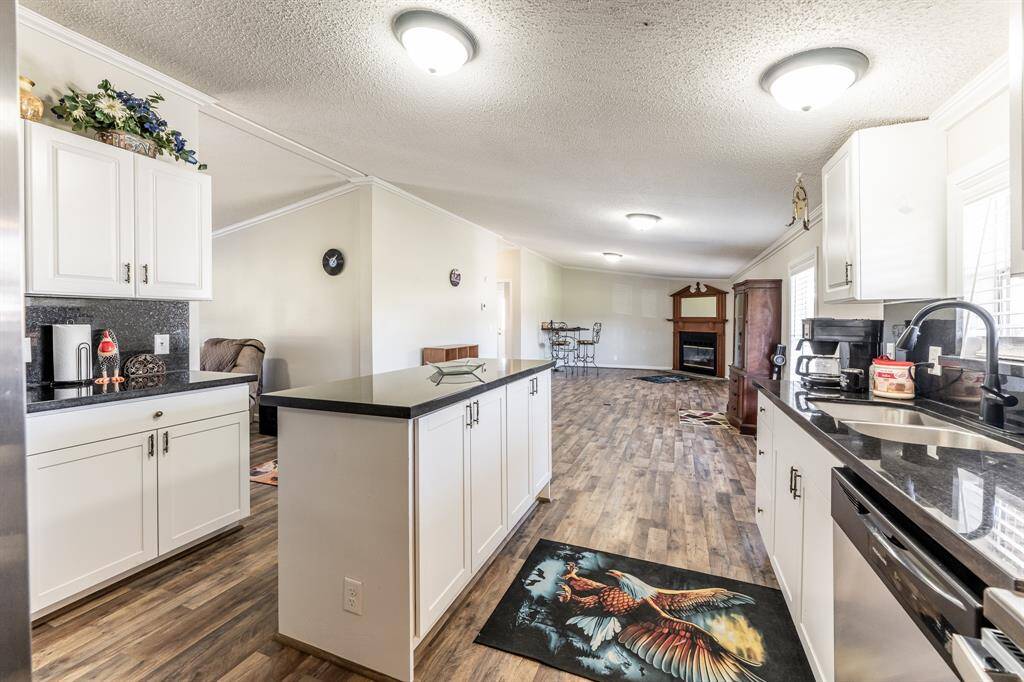 This bright, open-concept kitchen and living area features modern white cabinetry, sleek black countertops, and hardwood flooring. The space extends to a cozy living room with a fireplace, creating a warm and inviting atmosphere.