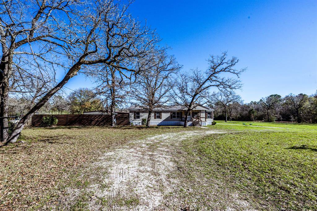 This photo shows a charming, single-story home set on a spacious, grassy lot with mature trees. A gravel driveway leads up to the house, offering a sense of privacy and tranquility. Ideal for those seeking a rural setting.